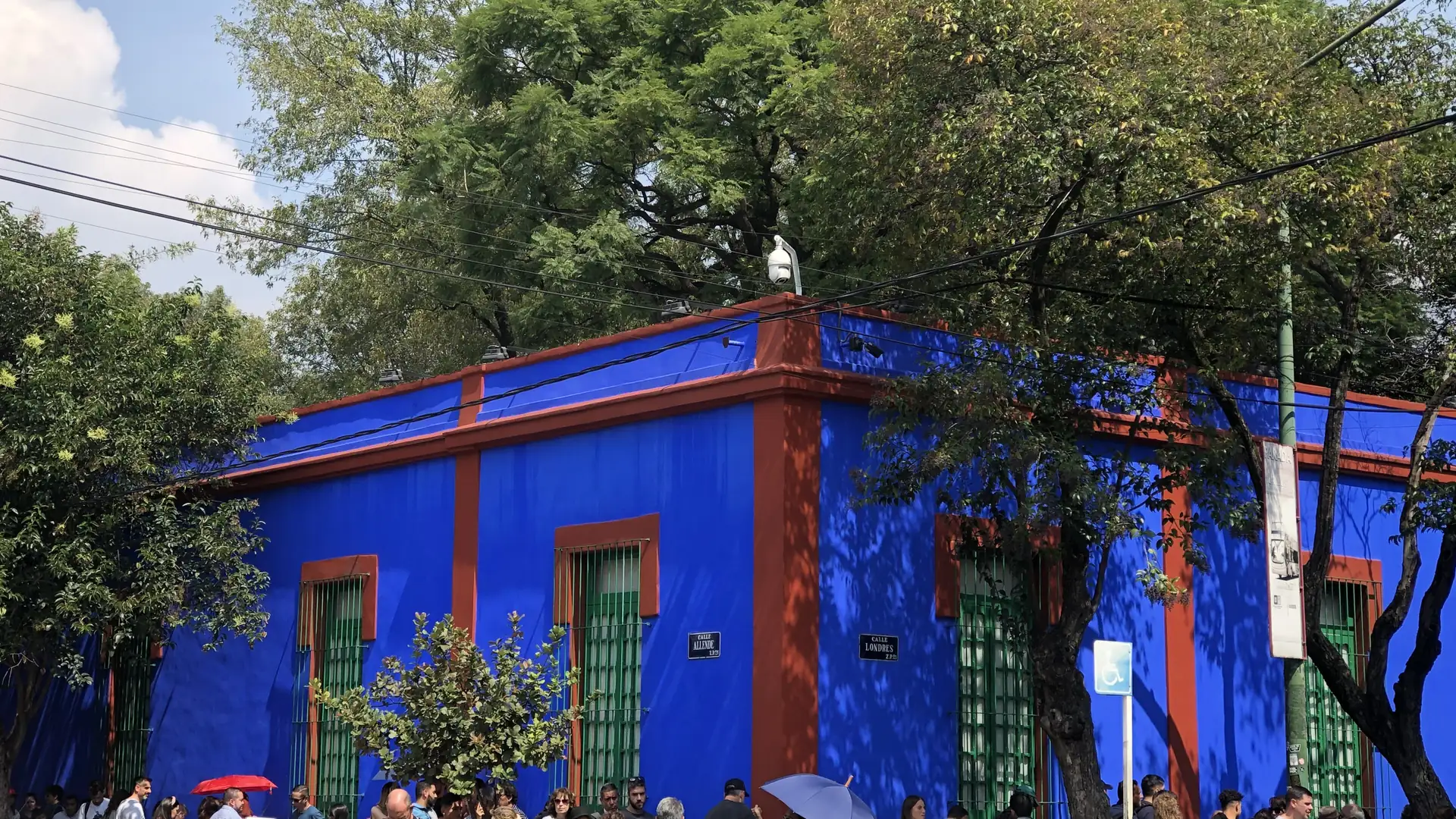 Nároží Casa Azul v Coyoacánu: sytě modré zdi s červenohnědým lemem a vysoká okna s zelenými mřížemi, pod mohutnými stromy a modrým nebem, zatímco návštěvníci stojí ve frontě podél chodníku.