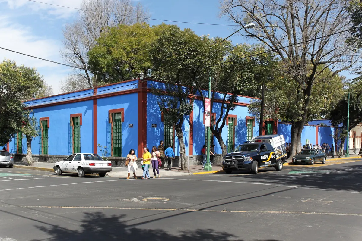 The long blue walls and red trim of the Casa Azul seen across a street junction in Coyoacán, under tall trees