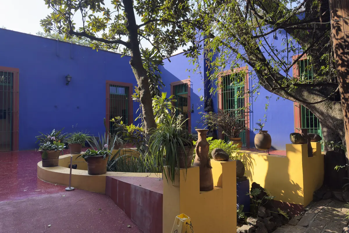 Nádvoří Casa Azul s tmavě modrými zdmi, zelenými rámy oken, květináči na žlutém vyvýšeném záhonu a červeně natřenou podlahou pod velkým stromem.