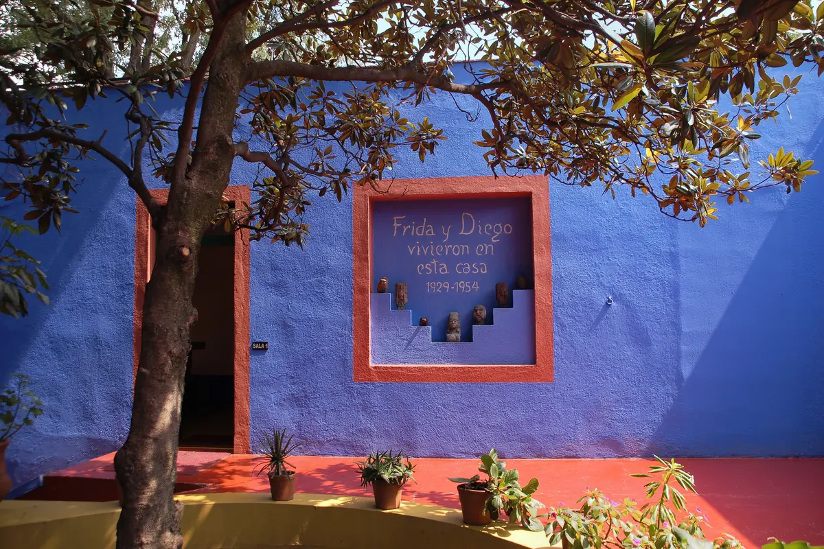 Modrá zeď nádvoří s červeným rámem, v němž stojí namalovaná slova Frida y Diego vivieron en esta casa 1929–1954, nad drobnými kamennými figurkami vsazenými do stupňovitých výklenků.
